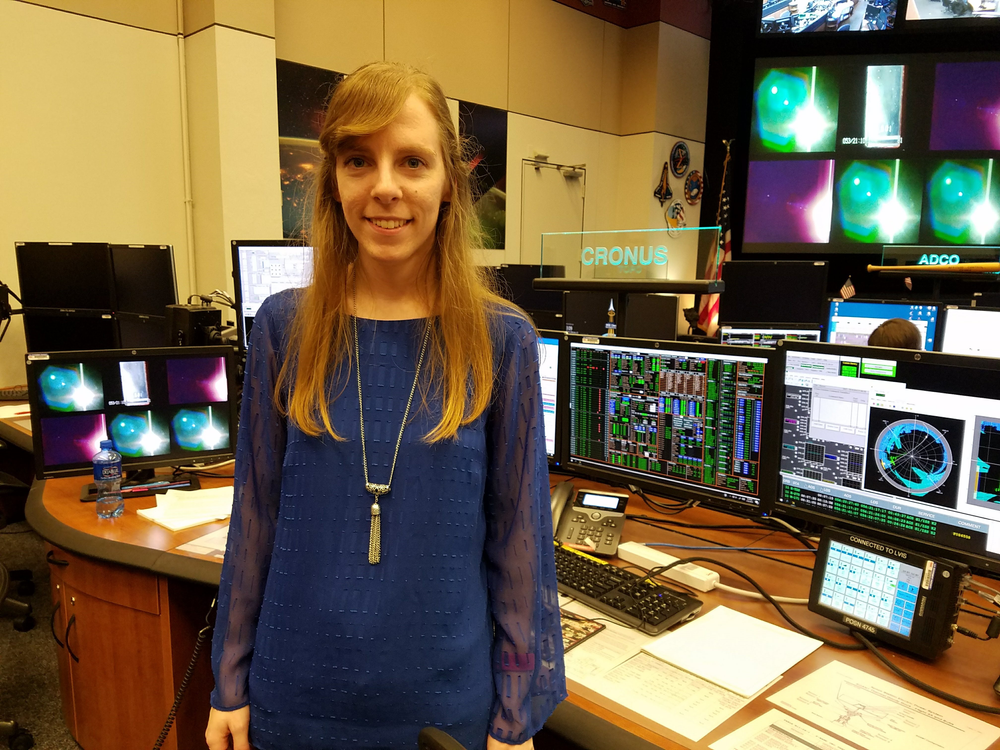 A young woman stands in front of a desk with multiple computer and TV screens behind her.